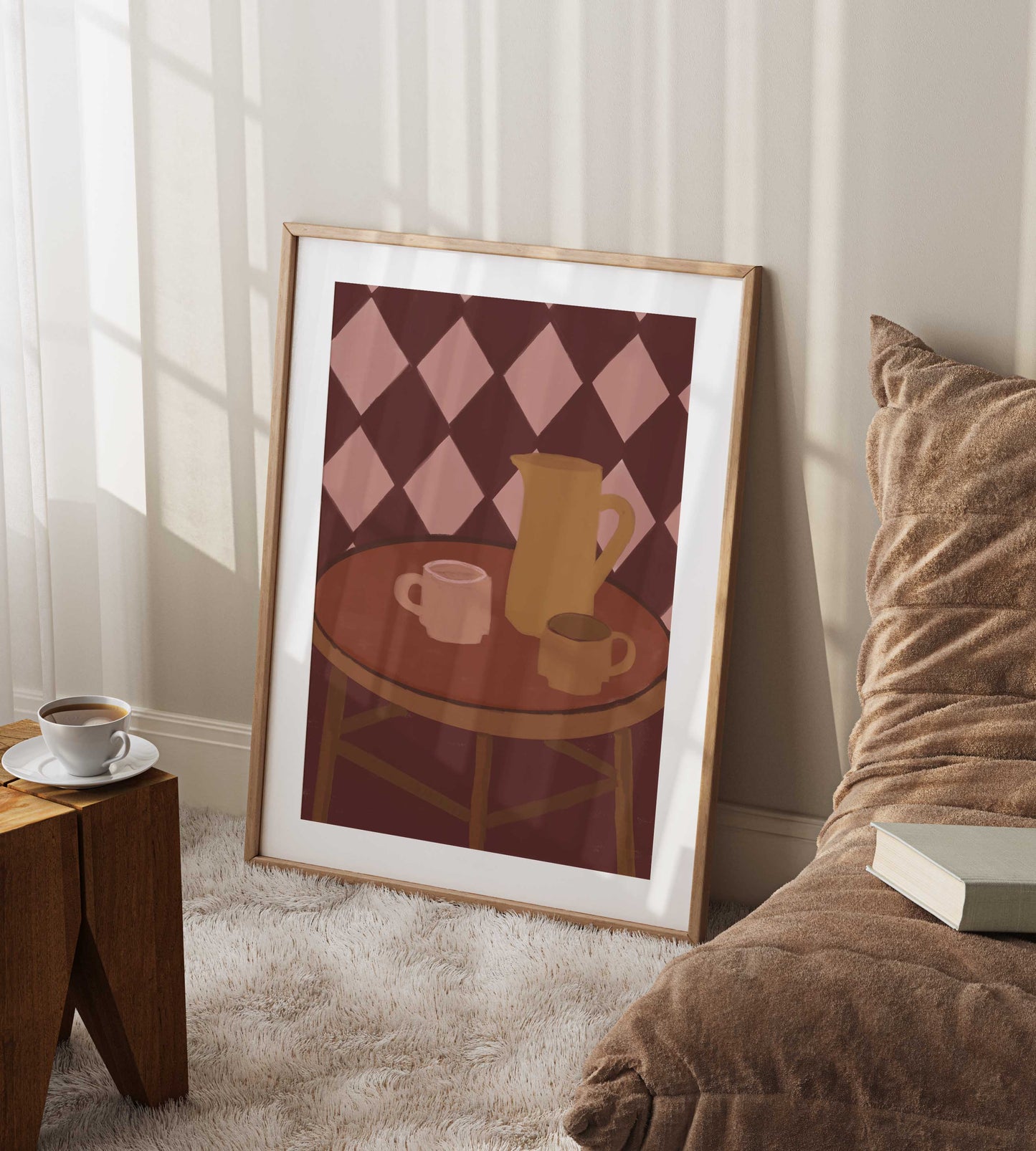 A framed art print leaning against a wall in a cozy living room setting. The print features a minimalist illustration of a round wooden table with a yellow pitcher and two pink mugs, set against a pink and brown diamond patterned background.