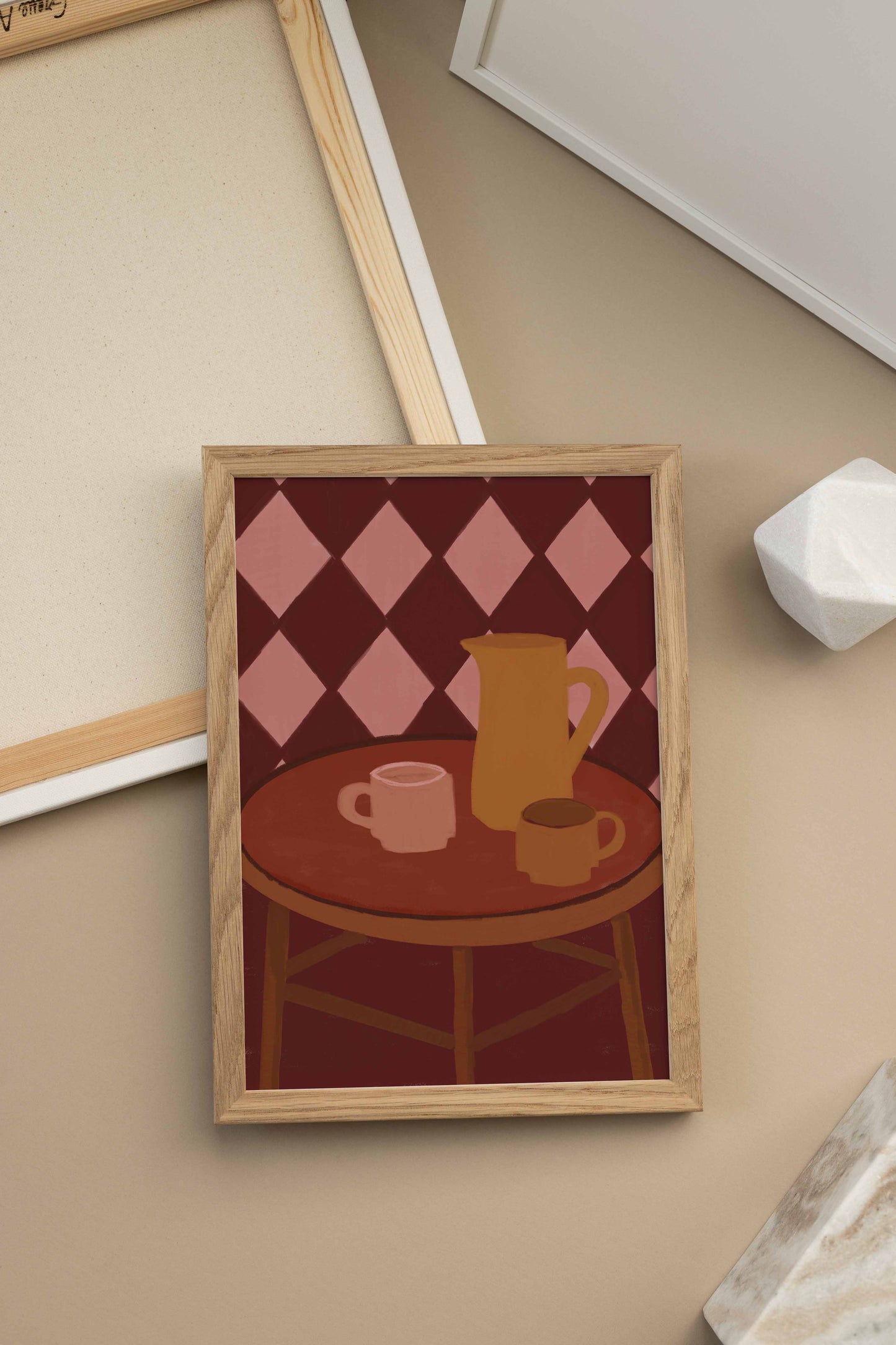 A framed art print featuring a stylized illustration of a round wooden table with a pink and burgundy diamond-patterned background, a yellow pitcher, and two pink cups, displayed on a beige surface.