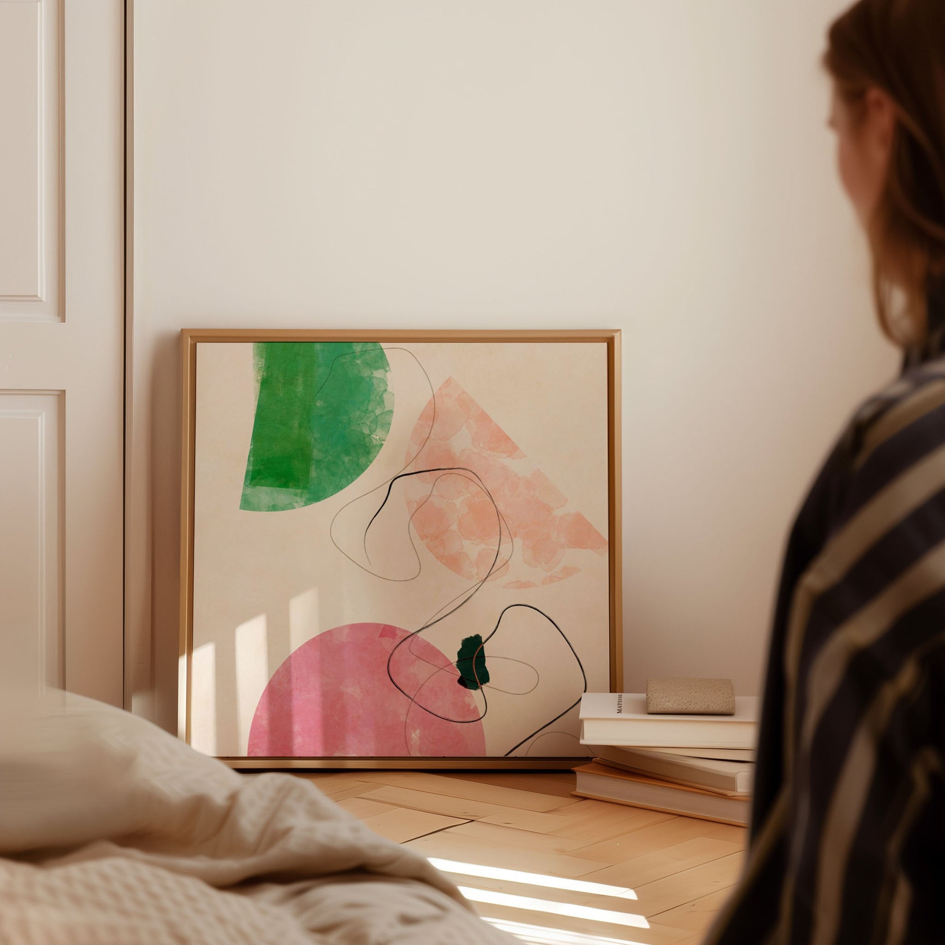 A framed abstract art print with green, pink, and peach shapes and black line art, leaning against a cream wall in a softly lit bedroom. A person in a striped shirt is partially visible in the foreground, and books and a bed are in the room.