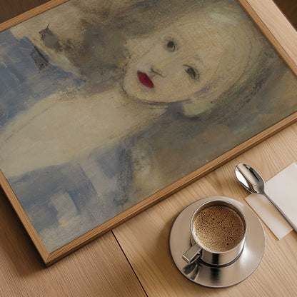 A framed art print featuring an impressionistic portrait of a woman with pale skin and red lips, displayed on a light wood table next to a cup of coffee, a spoon, and a napkin.
