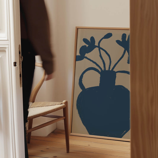 A framed minimalist art print featuring a dark blue vase with flowers on a light beige background, leaning against a wall in a warm, modern room next to a wooden chair.