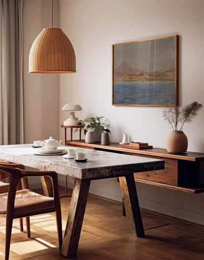A styled dining room corner featuring a rustic wooden table with a stone-look top, a woven pendant light, a wall-mounted console shelf with plants and a framed landscape painting, and a wooden chair in a warm neutral interior.