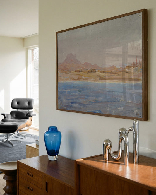 A framed landscape painting hanging on a beige wall in a modern room, featuring a blue sea and brown mountains, with a wooden console table and a blue glass vase below.