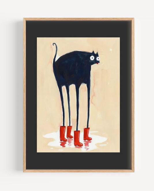 Framed wall art featuring a stylized black cat with long legs and red rain boots on a cream background, shown in a light wood frame with a black mat.