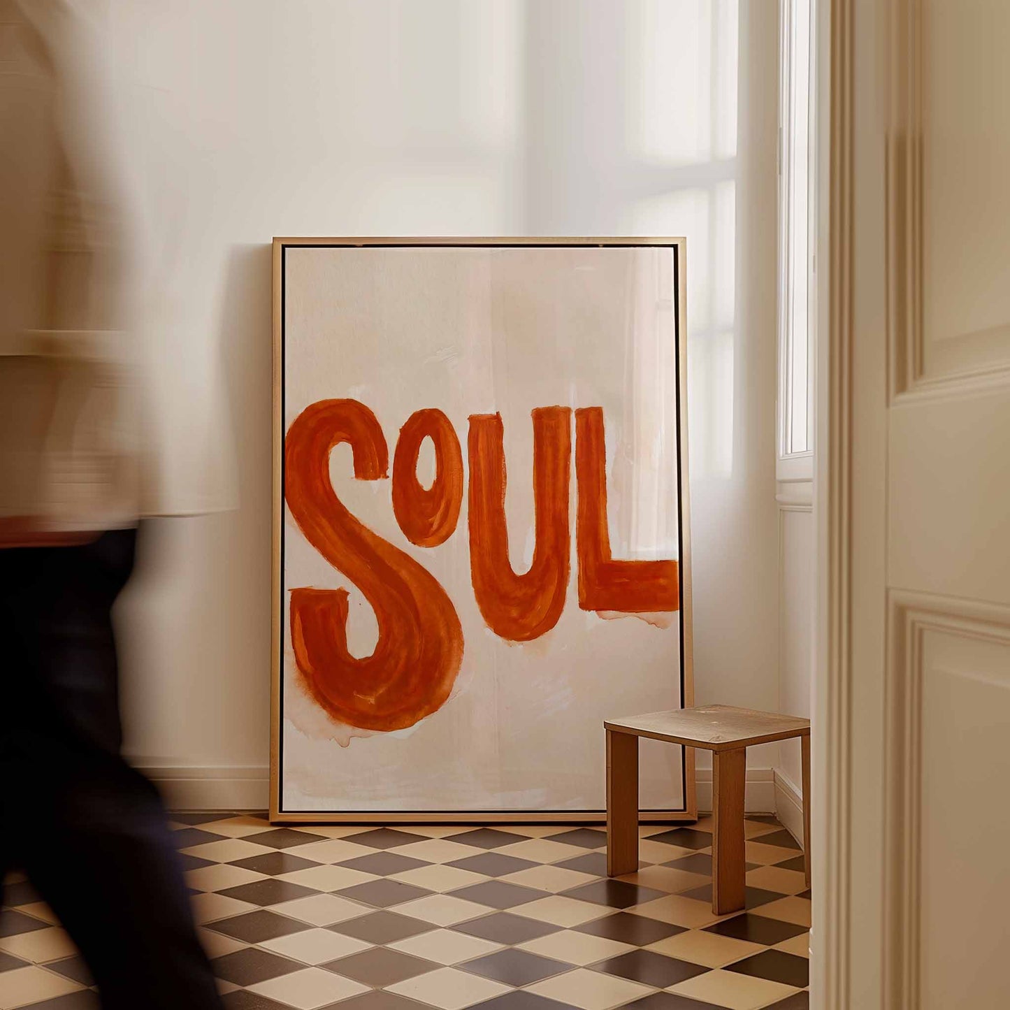 A large framed art print featuring the word 'SOUL' in bold orange-red brushstrokes on a cream background, leaning against a white wall in a room with a checkered floor and a small wooden stool.