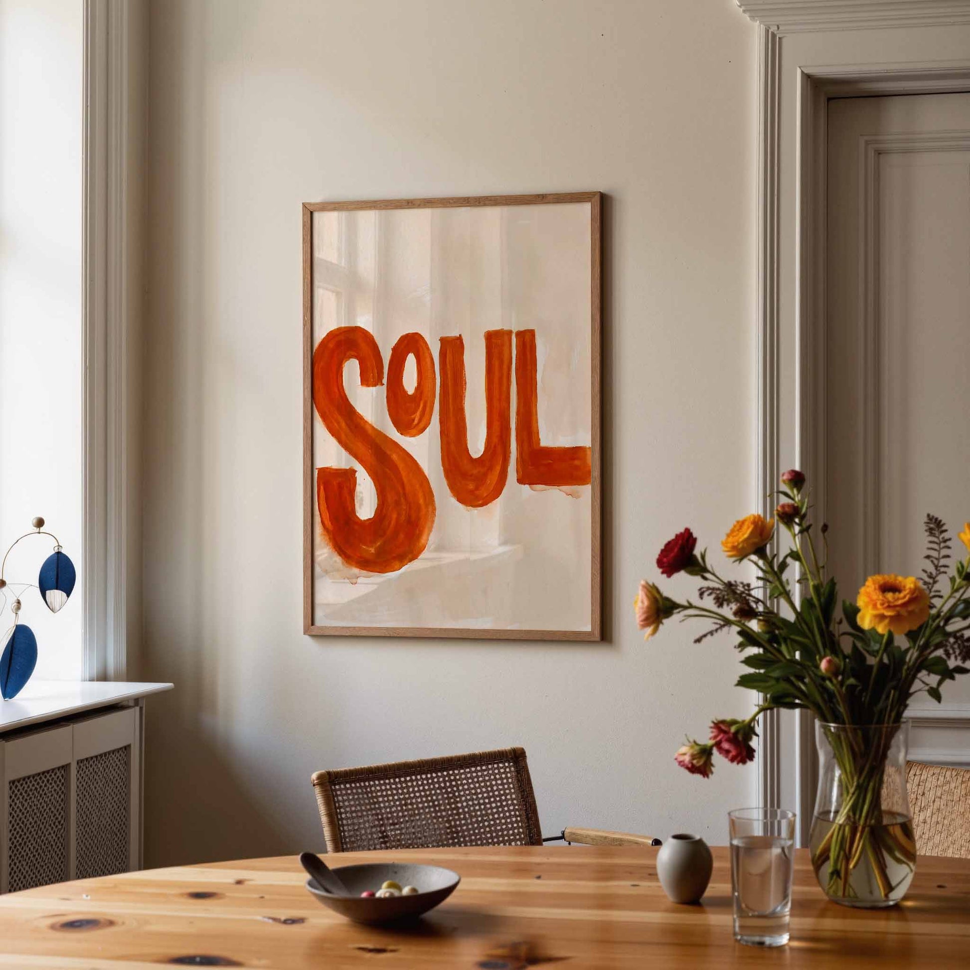 Framed wall art with the word “SOUL” in large orange letters on a cream background, hanging above a wooden dining table in a styled interior with flowers and a chair.