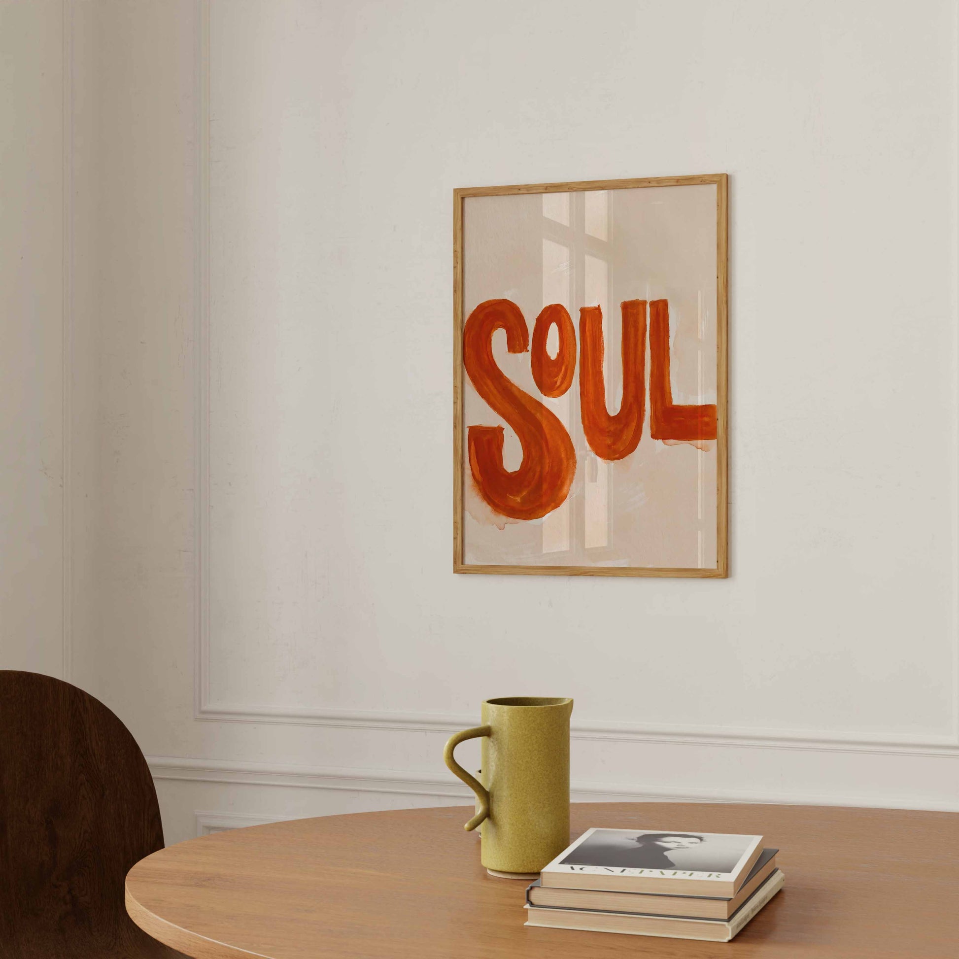 Framed wall art with the word “SOUL” in large orange-red letters on a light beige background, hanging on a white wall above a wooden table with a green mug and stacked books.