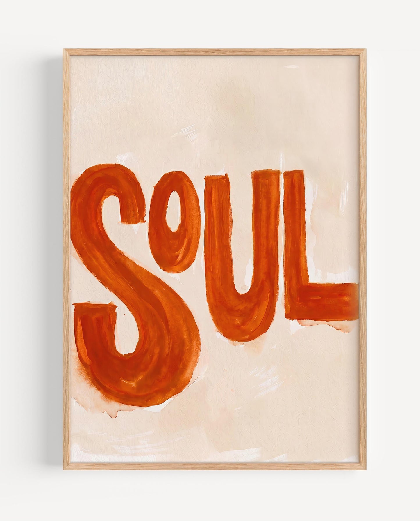 Framed wall art featuring the word “SOUL” painted in bold orange-red lettering on a warm cream background, shown in a light natural wood frame against a white wall.