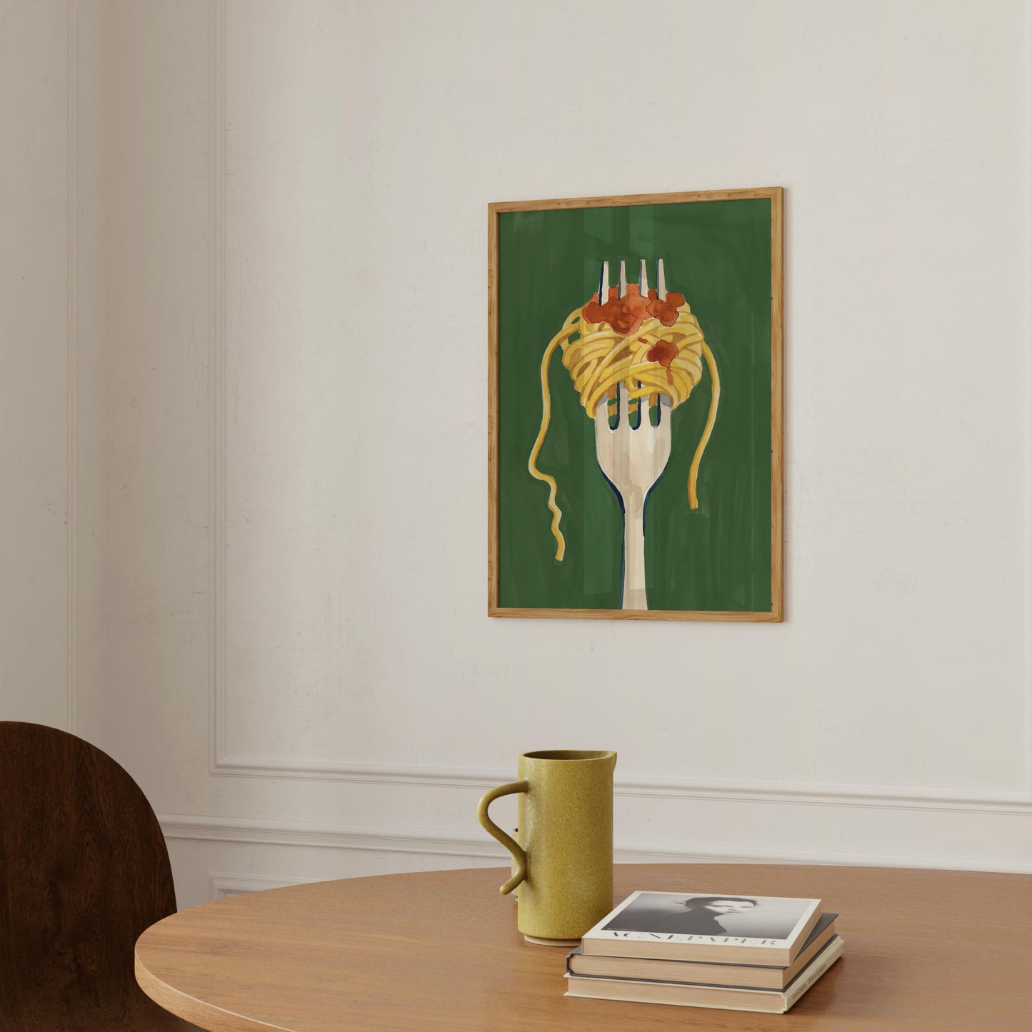 A framed art print depicting spaghetti on a fork over a dark green background, hanging on a white wall above a wooden table with a yellow-green mug and a stack of books.