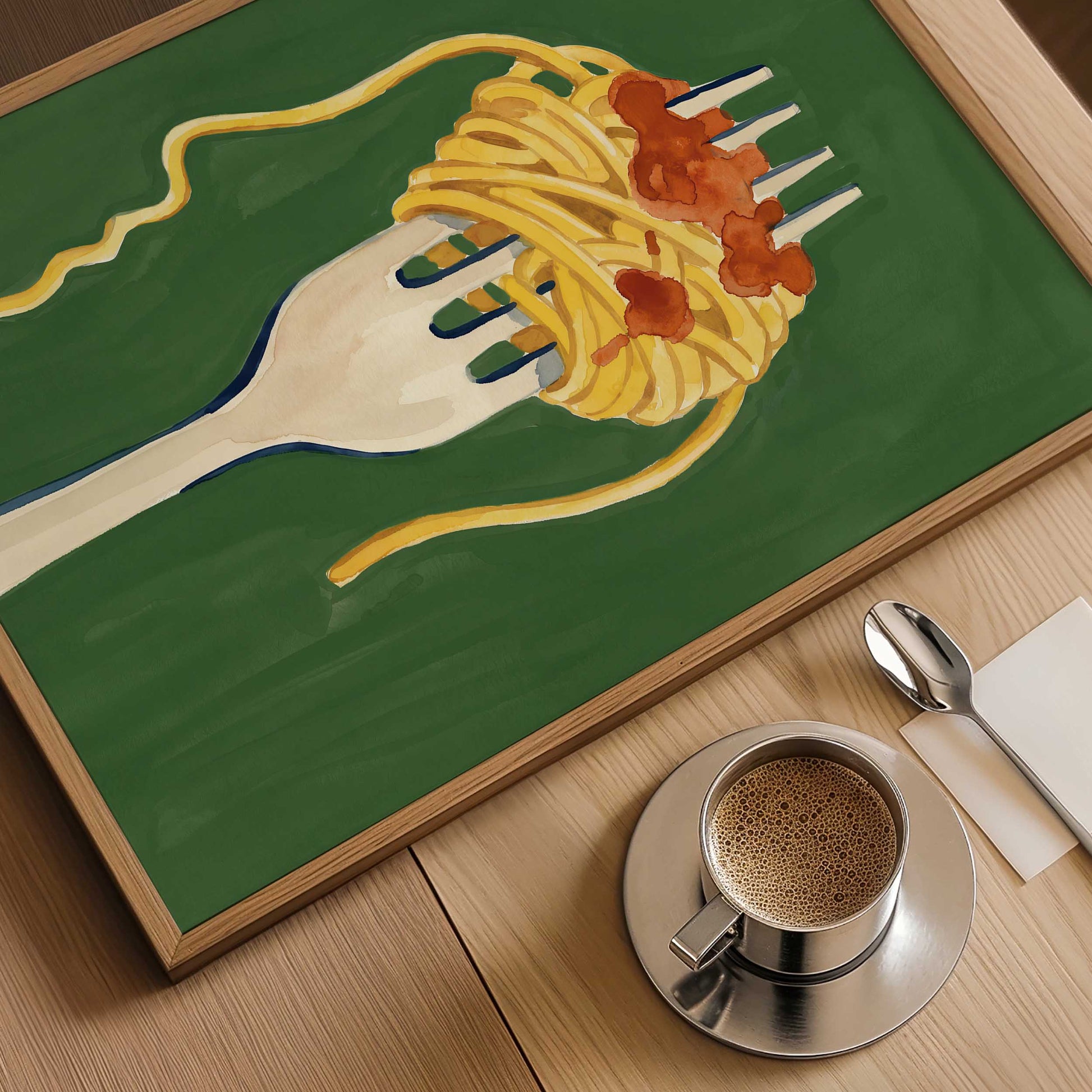 A framed art print depicting a white fork twirling spaghetti with red tomato sauce on a green background, styled on a wooden table next to a cup of coffee, a spoon, and a napkin.