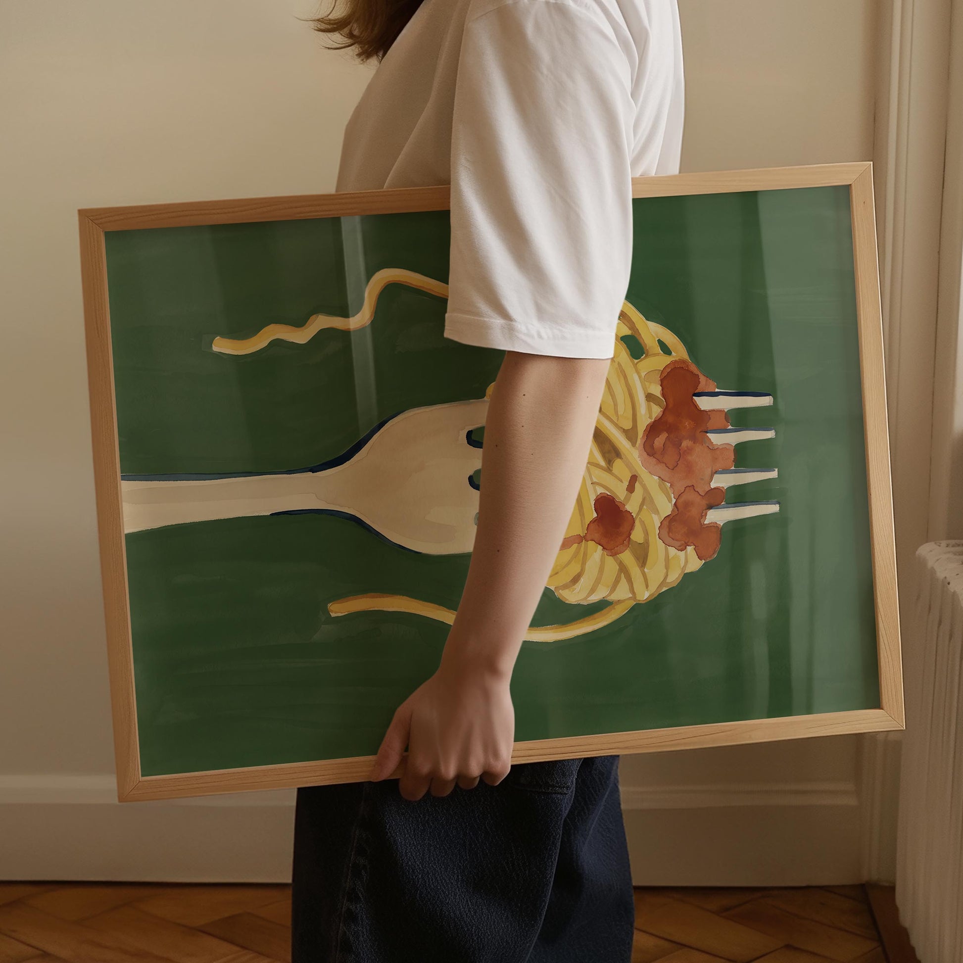 A person in a white t-shirt and dark pants holds a framed art print featuring a whimsical illustration of spaghetti and meatballs on a fork against a dark green background.