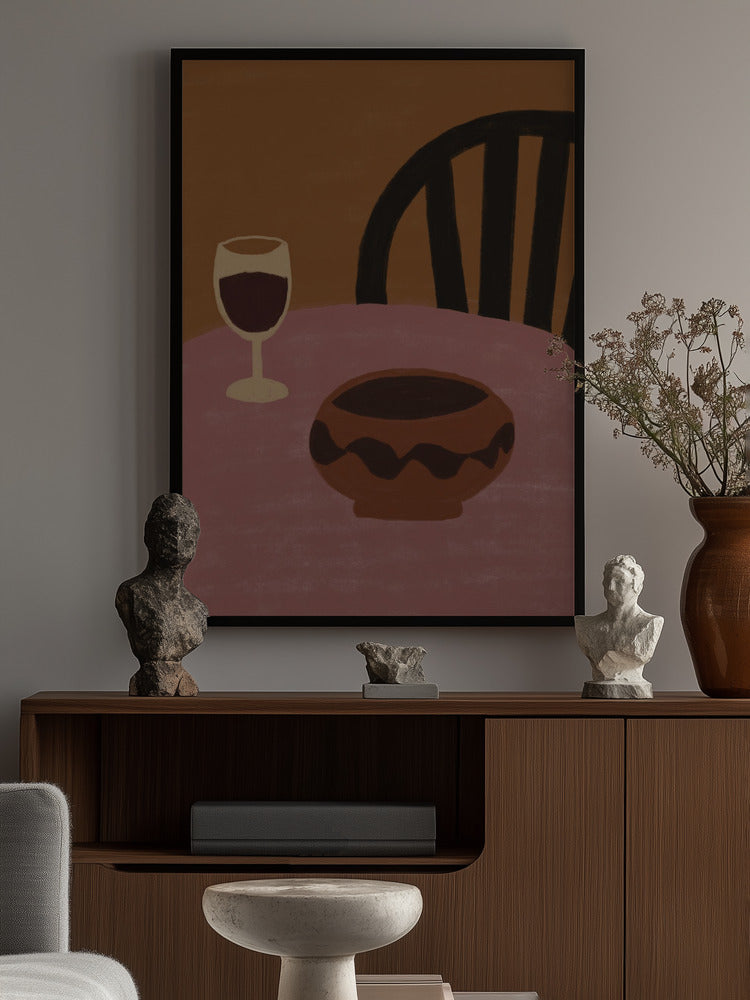 A framed abstract art print featuring a wine glass, a bowl, and a chair in muted brown, pink, and black tones, displayed on a wall above a wooden console table with decorative busts and a vase.