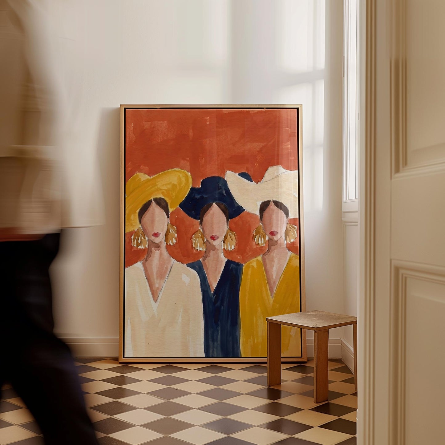 A large framed art print depicting three stylized women in colorful clothing, leaning against a white wall in a room with a checkered floor and a small wooden stool.