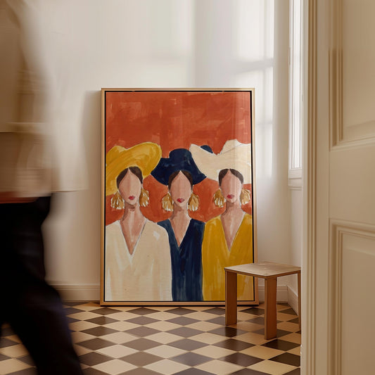 A large framed art print depicting three stylized women in colorful clothing, leaning against a white wall in a room with a checkered floor and a small wooden stool.