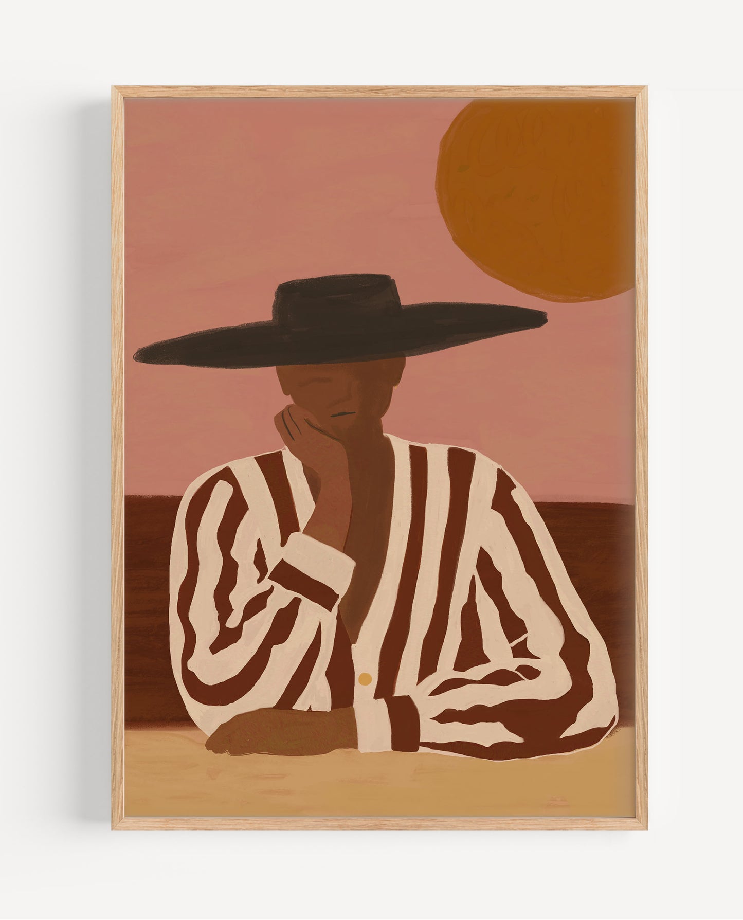 A framed art print featuring a stylized illustration of a person in a black wide-brim hat and brown-and-cream striped shirt, seated against a dusty pink sky with a large orange sun, shown against a light gray wall.