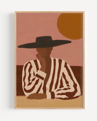 A framed art print featuring a stylized illustration of a person in a black wide-brim hat and brown-and-cream striped shirt, seated against a dusty pink sky with a large orange sun, shown against a light gray wall.