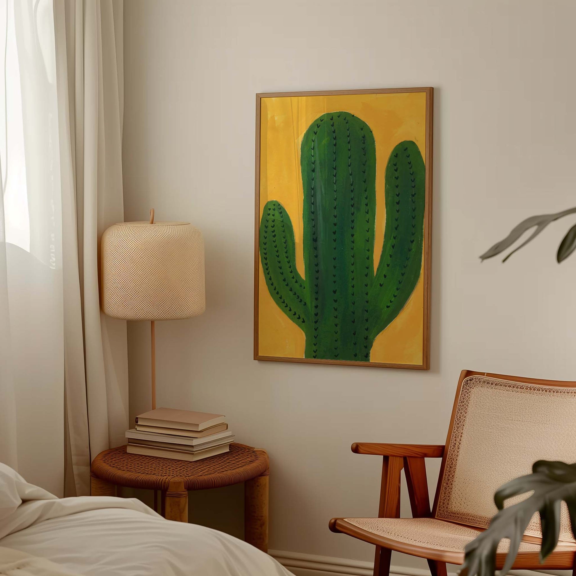 A framed vertical art print featuring a green cactus on a mustard yellow background, displayed on a light beige wall in a styled bedroom setting next to a lamp, books, and a chair.