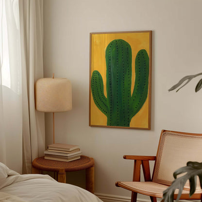 A framed vertical art print featuring a green cactus on a mustard yellow background, displayed on a light beige wall in a styled bedroom setting next to a lamp, books, and a chair.
