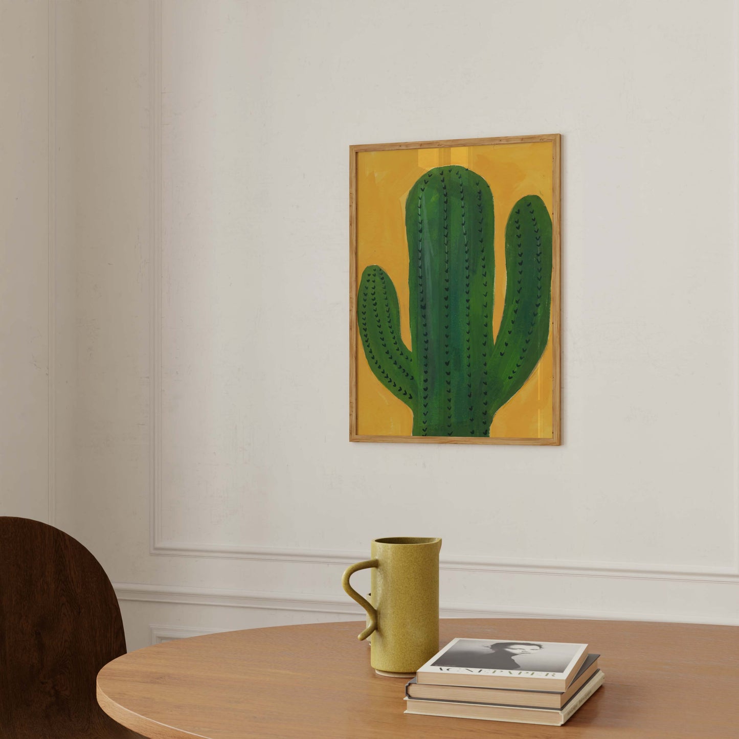 A framed art print of a green cactus on a mustard yellow background, hanging on a white wall above a wooden table with a green ceramic mug and a stack of books.