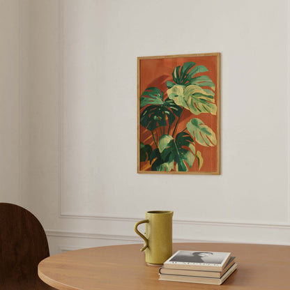 A framed botanical art print featuring green monstera leaves on a terracotta background, hanging on a white wall above a wooden table with a yellow-green ceramic mug and a stack of books.