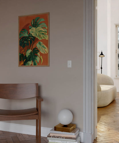 A framed botanical art print featuring green monstera leaves on an orange background, hanging on a beige wall in a modern interior setting with a wooden bench and a white globe lamp.