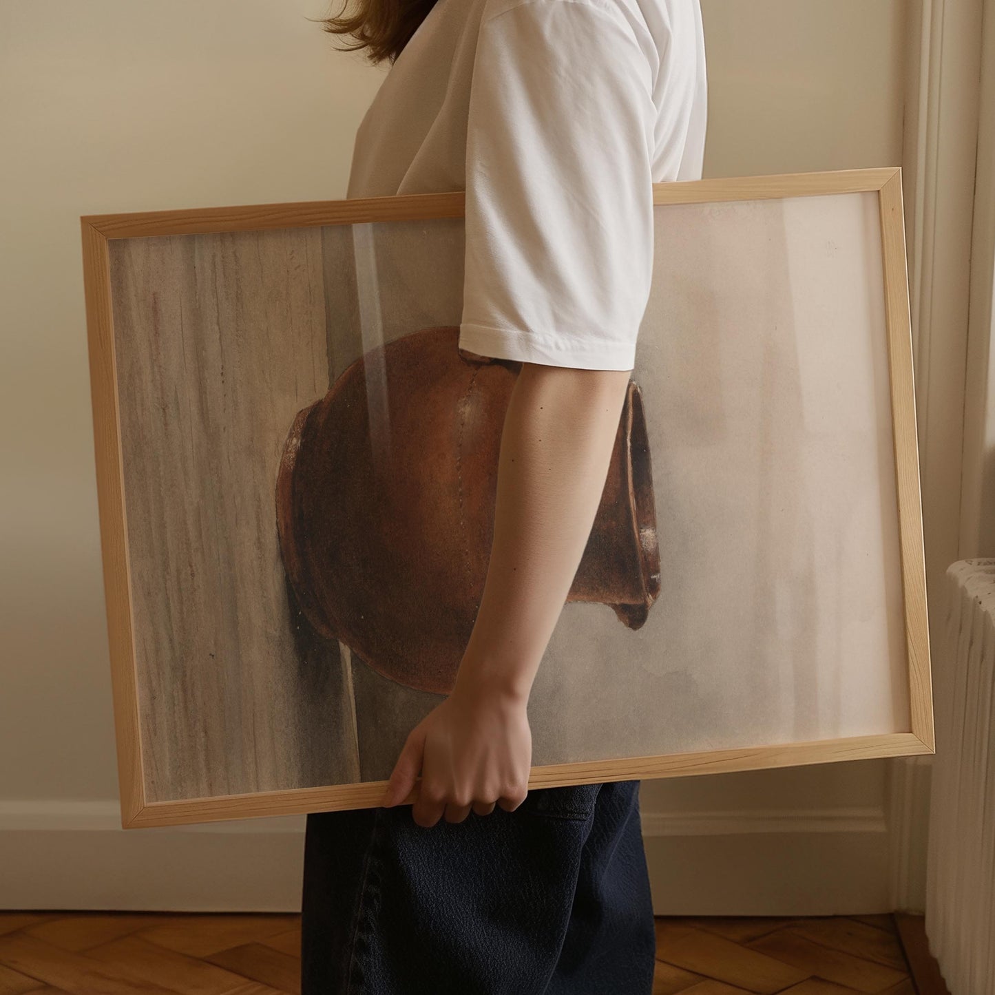 A person in a white t-shirt and dark jeans holds a large framed art print. The print features a minimalist still life of a brown ceramic pot on a light beige background, enclosed in a light natural wood frame.