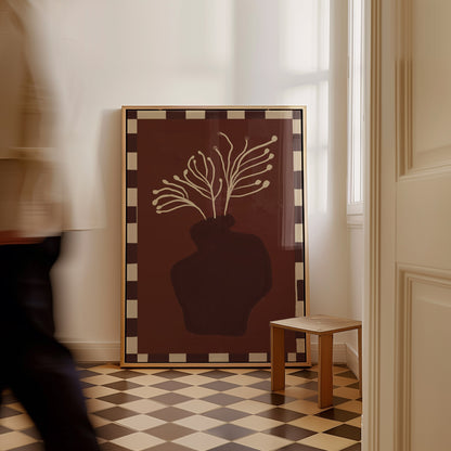 A large framed art print leaning against a white wall in a room with a checkered floor. The print features a dark brown background with a cream-colored silhouette of a vase holding branching branches, bordered by a cream and dark brown checkered frame.