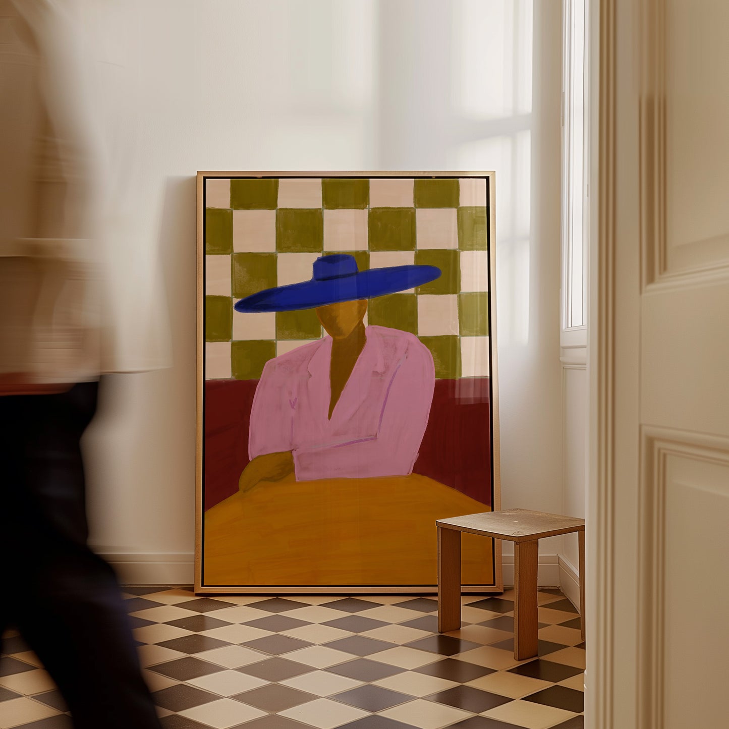 A framed art print depicting a stylized figure in a pink shirt and blue hat against a green and beige checkerboard background, leaning against a white wall in a room with a checkered floor and a small wooden stool.