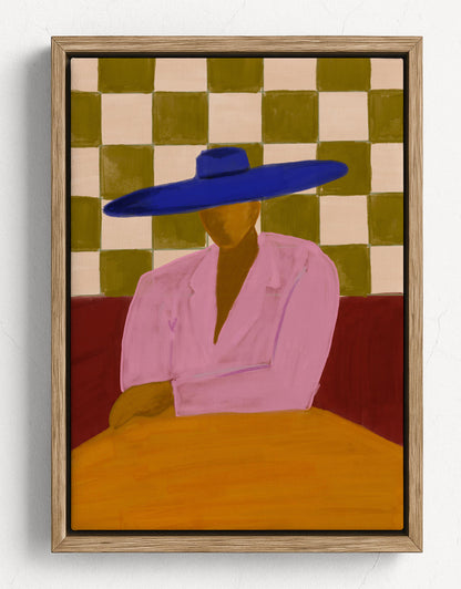 A framed art print featuring a stylized figure in a pink shirt and wide blue hat seated at a yellow table, with a green and cream checkered background, shown in a light wood frame against a white wall.