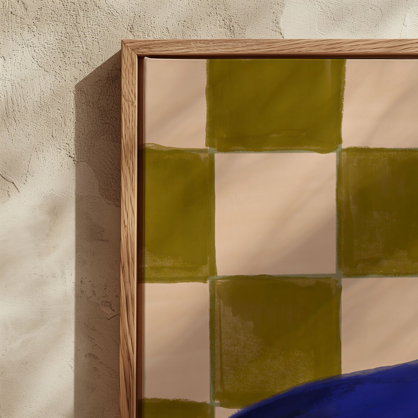 A close-up of a framed art print featuring a checkered pattern of olive green and cream squares, set against a textured beige wall with soft shadows.