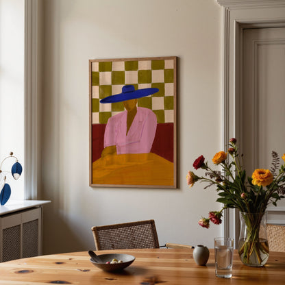A framed art print featuring a stylized figure in a pink shirt and blue hat against a green and beige checkered background, hanging on a light beige wall in a dining room setting with a wooden table and flowers.