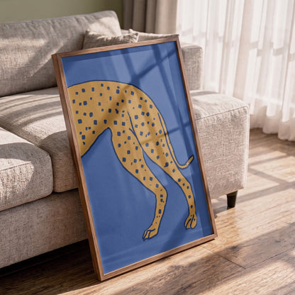 A framed art print featuring a stylized yellow cheetah with blue spots on a blue background, leaning against a light gray sofa on a wooden floor in a sunlit living room.