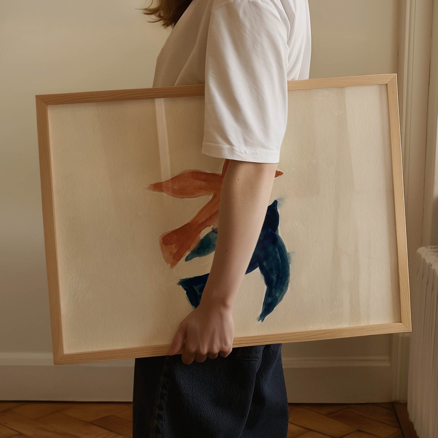 A person in a white t-shirt and dark skirt holds a large framed abstract painting featuring blue and orange brushstrokes on a cream background with a light wood frame.