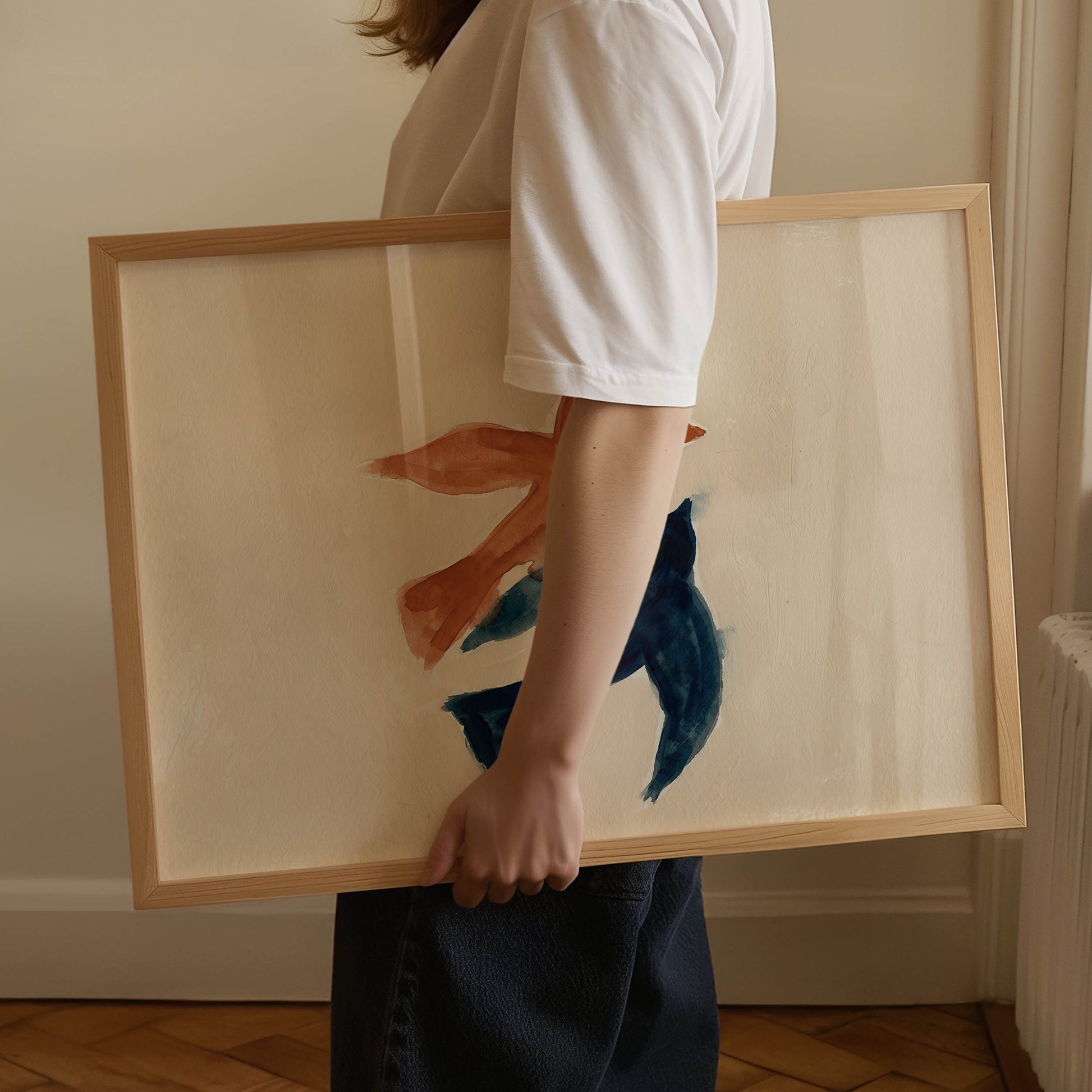 A person in a white t-shirt and dark skirt holds a large framed abstract painting featuring blue and orange brushstrokes on a cream background with a light wood frame.