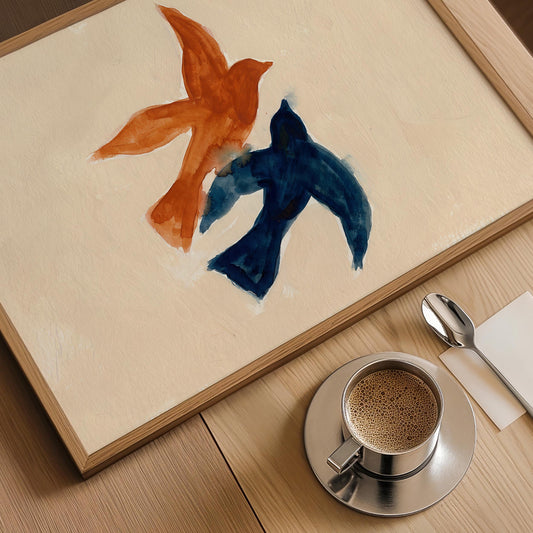 A framed abstract painting featuring two bird-like forms in orange and dark blue on a cream background, displayed on a wooden table next to a cup of coffee, a spoon, and a napkin.