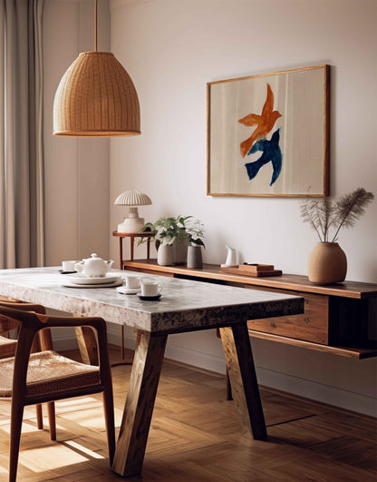 A styled interior scene showing a dining area with a wooden table, chairs, pendant light, wall console, and framed abstract artwork on a beige wall.
