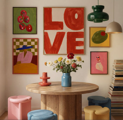 A styled interior scene with multiple colorful framed wall art pieces above a round wooden table. The artwork includes abstract lettering, fruit, a figure in a hat, and a pink figure, while the room also includes hanging pendant lights, a blue vase with flowers, colorful stools, and a stack of books.