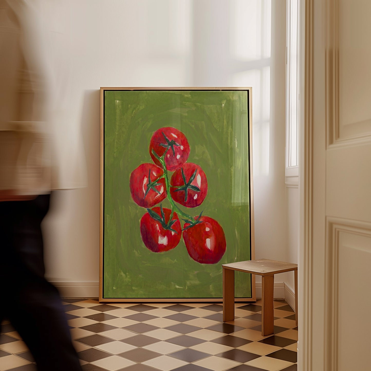 A framed painting of five red tomatoes on a green background, leaning against a white wall in a room with a checkered floor and a small wooden stool. A blurred person walks past on the left.