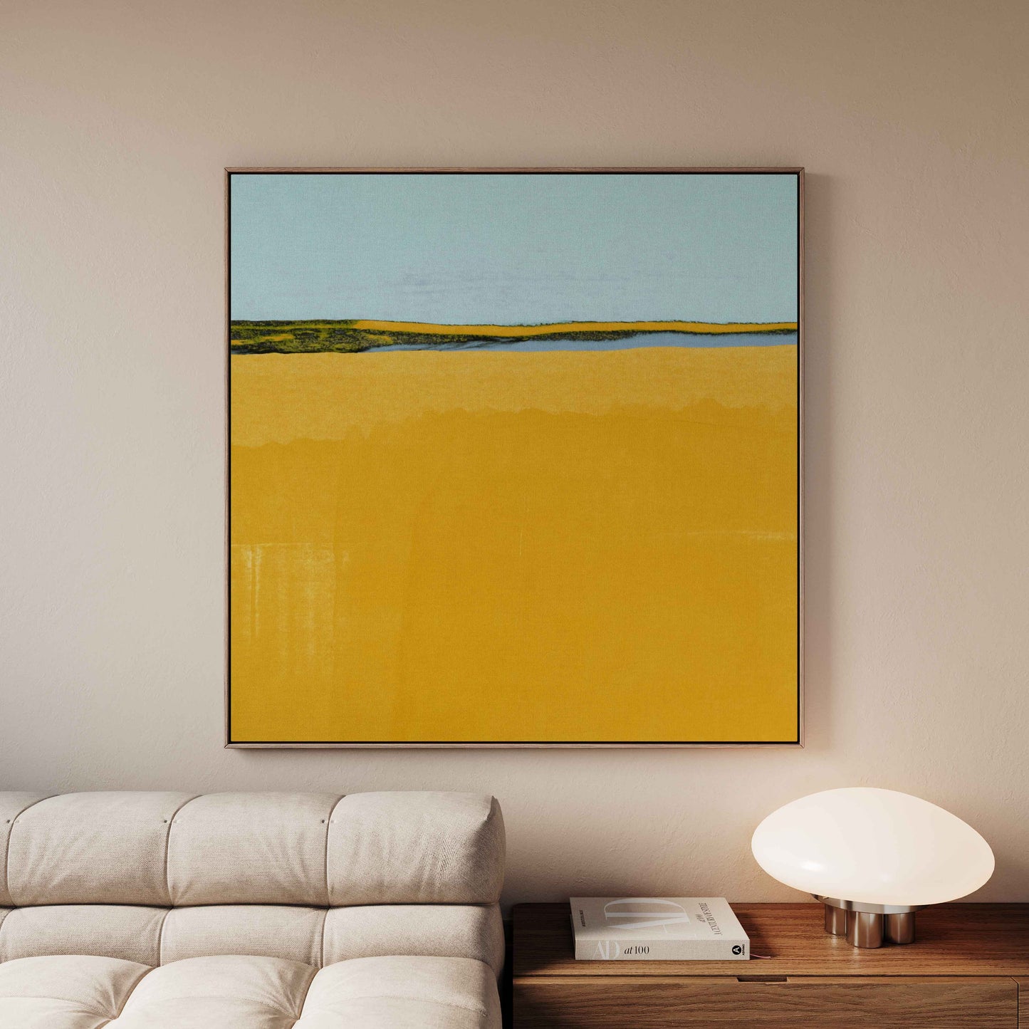A framed abstract landscape painting featuring a large yellow field and a light blue sky, displayed on a beige wall above a modern sofa and side table.