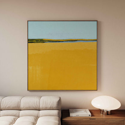 A framed abstract landscape painting featuring a large yellow field and a light blue sky, displayed on a beige wall above a modern sofa and side table.