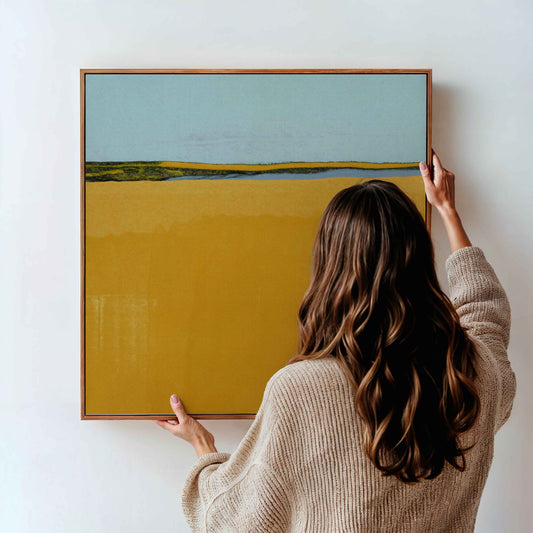 A woman in a beige knit sweater holds a framed landscape painting against a white wall. The painting features a large mustard yellow field under a pale blue sky with a thin green horizon line.