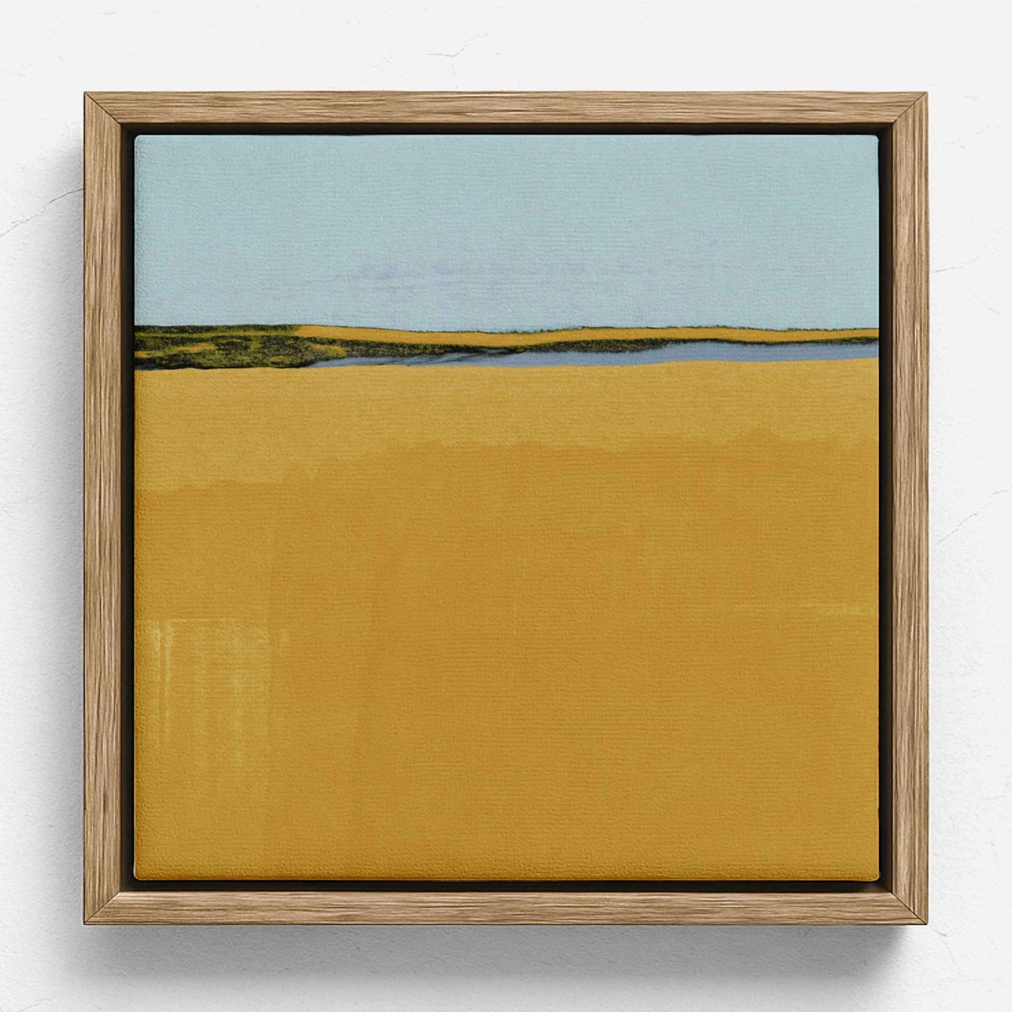 A square framed landscape painting featuring a wide mustard-yellow foreground, a distant strip of green and blue land, and a pale blue sky, set in a light natural wood frame against a white wall.