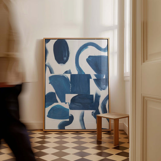 Large framed abstract art print with blue and white brushstrokes leaning against a cream wall in a room with a checkered floor and a small wooden stool. A blurred person walks past on the left.