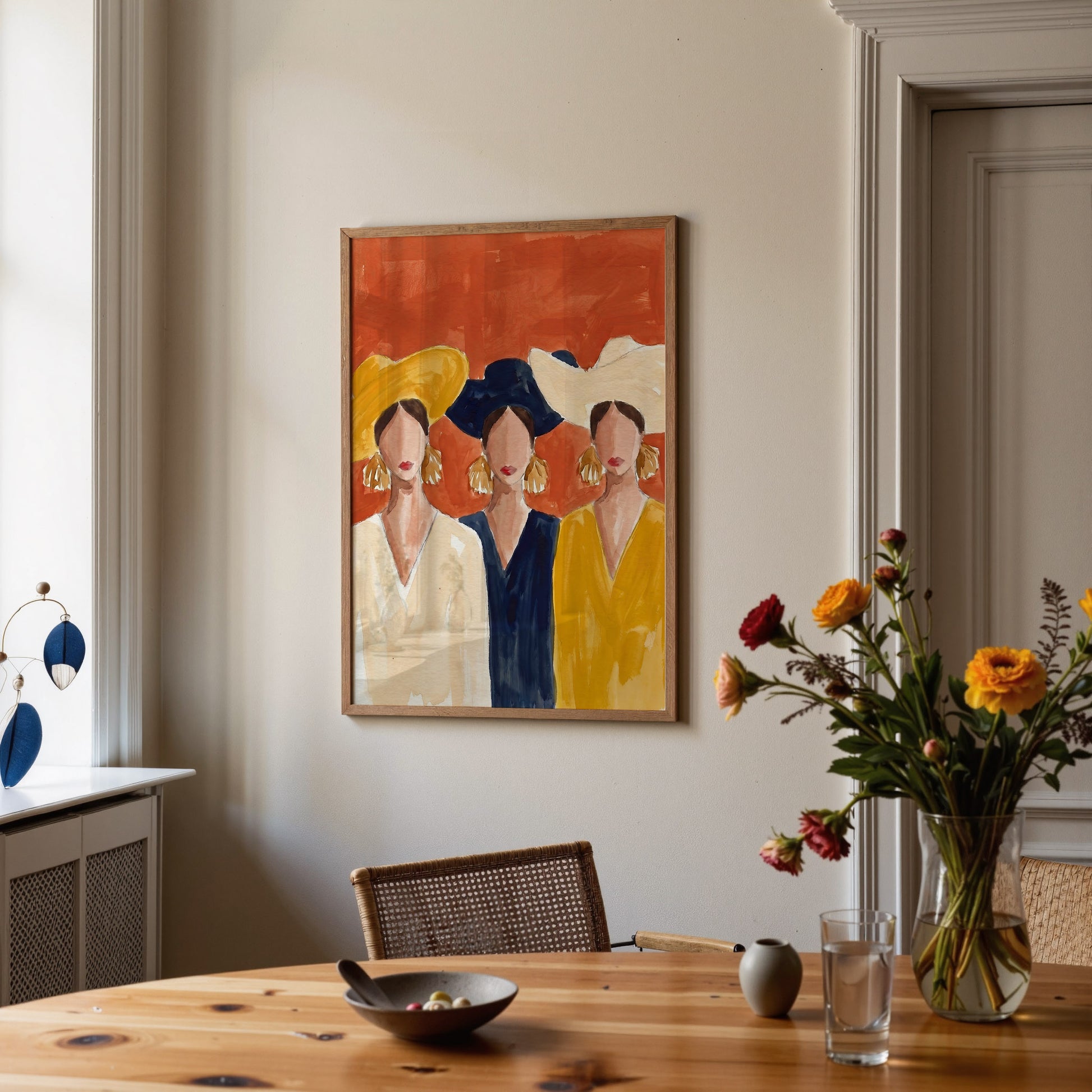 Dining room with a colorful abstract painting of three women on the wall, a wooden table with a vase of flowers, and a glass of water.