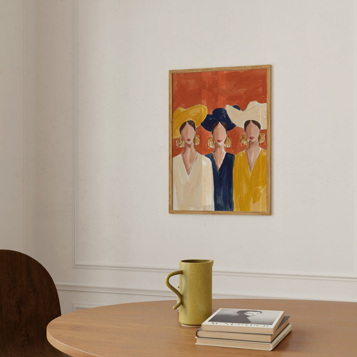 Framed artwork of three women on a wall above a table with a green mug and books.
